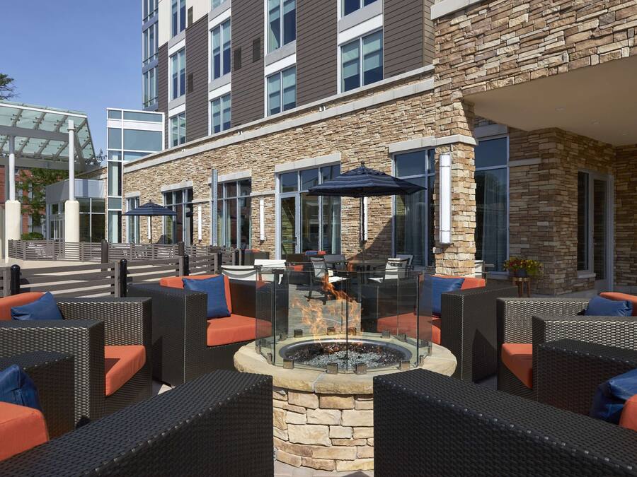 An outdoor seating area with wicker furniture and orange cushions, a fire pit, and a stone building with large windows.