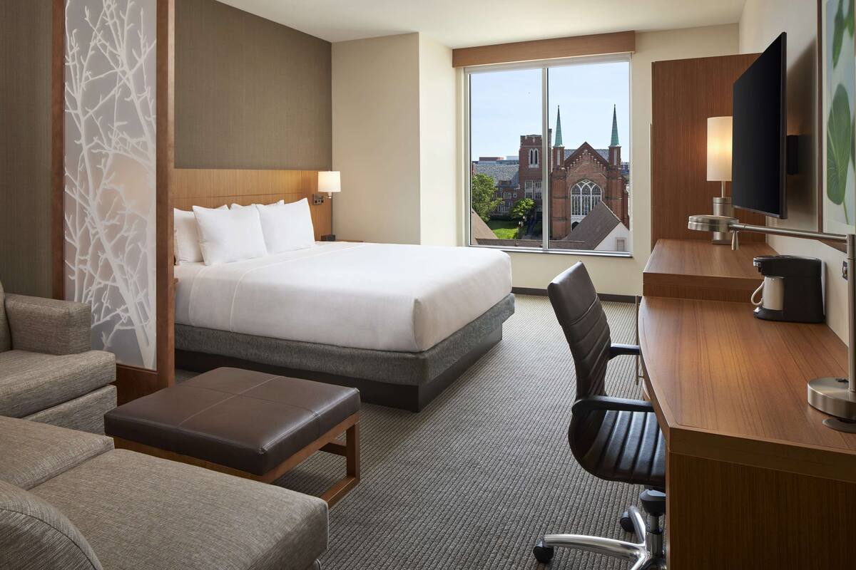 A hotel room with a large bed, a desk with a computer, a chair, and a view of a building with a spire.