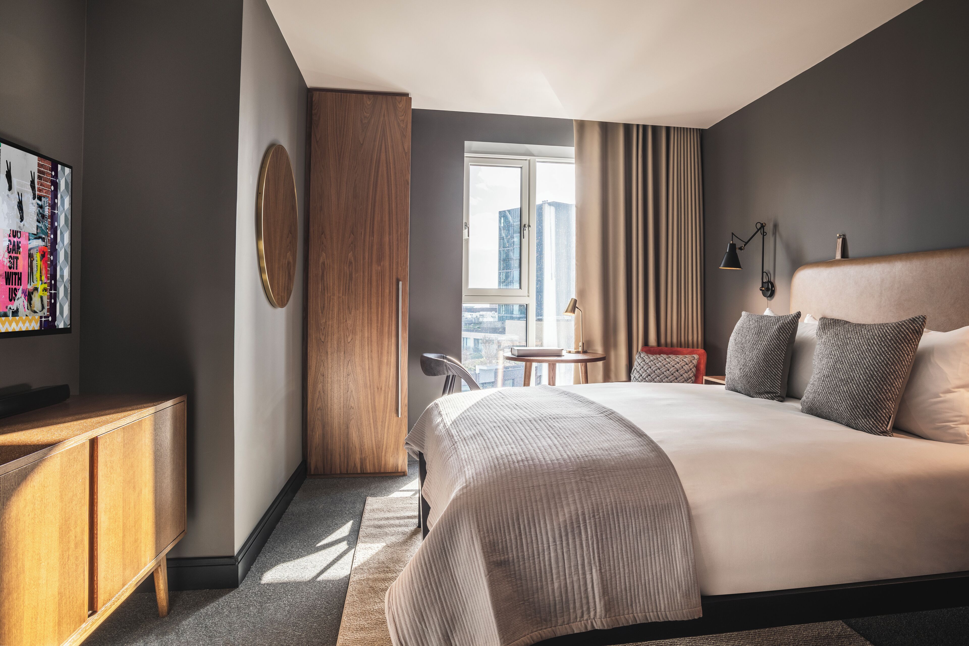 Ein modernes Hotelzimmer mit einem großen Bett, einem Holzschrank, einem runden Spiegel, einem kleinen Tisch und Blick auf die Stadt durch ein Fenster.