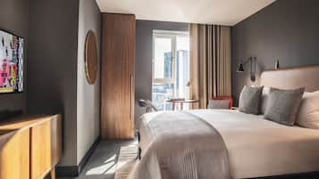 Ein modernes Hotelzimmer mit einem großen Bett, einem Holzschrank, einem runden Spiegel, einem kleinen Tisch und Blick auf die Stadt durch ein Fenster.