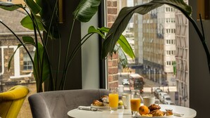 A dining area with a table set for breakfast, a view of the city through large windows, and a plant in the foreground. - The Gate London City (London)