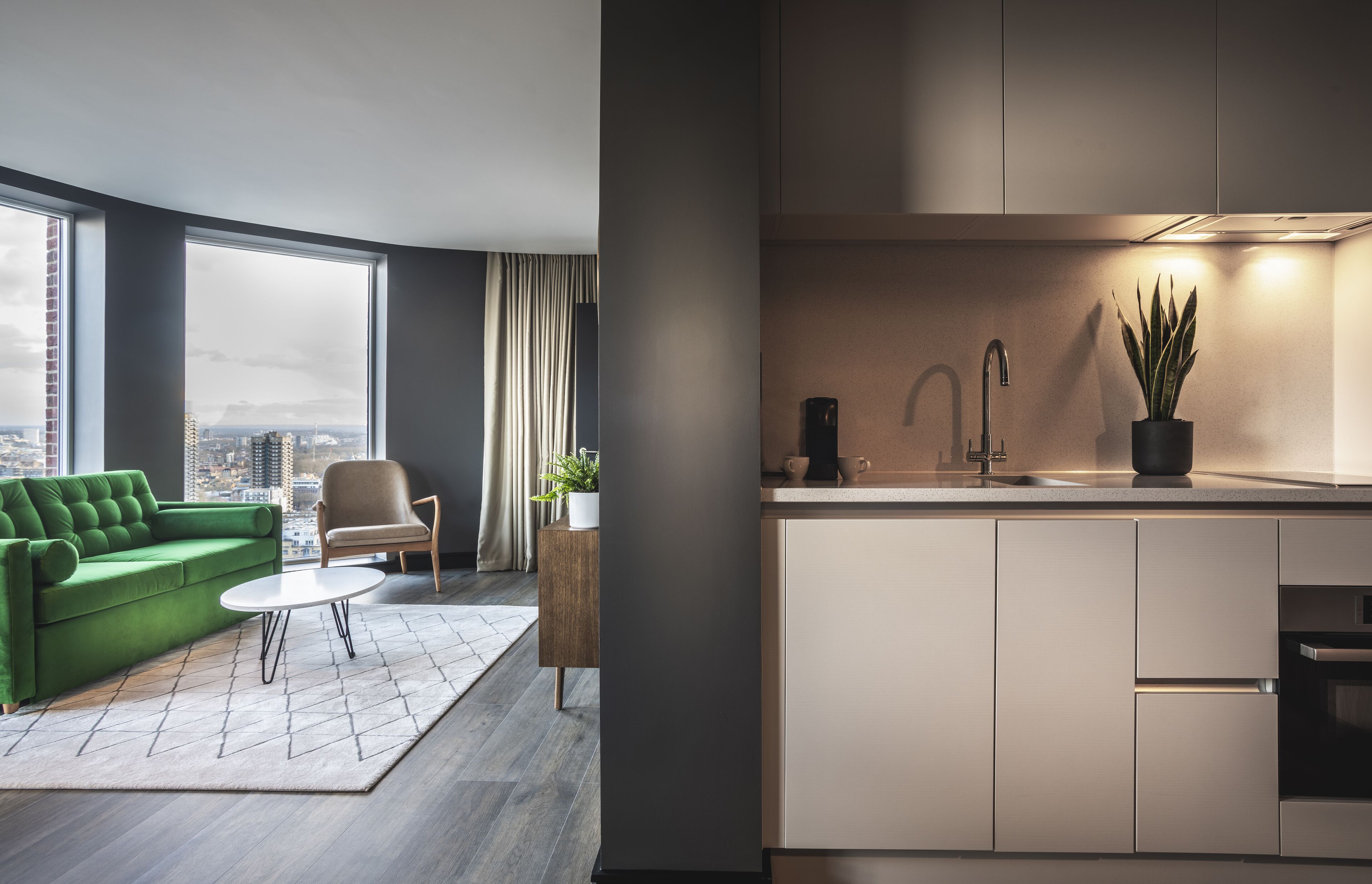 Ein modernes Wohnzimmer mit einem grünen Sofa, einem Couchtisch und Blick auf die Stadt durch große Fenster.
