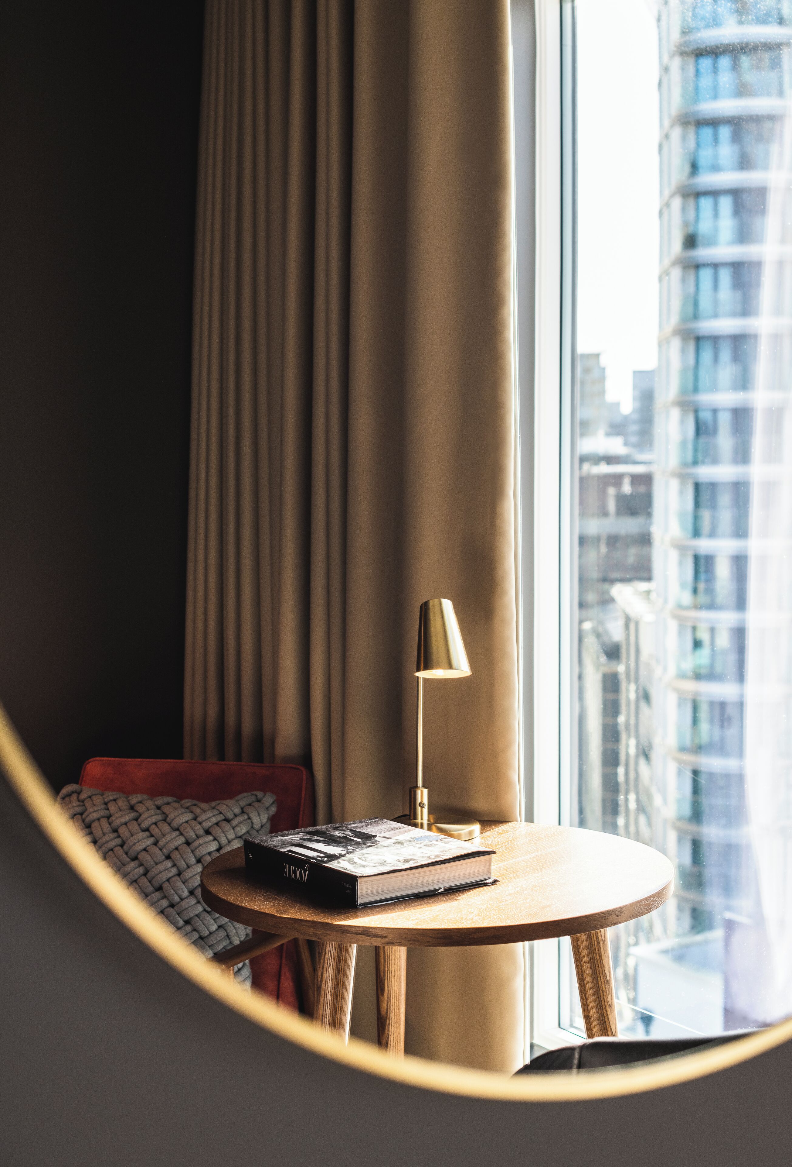 A round table with a book and a lamp, a window with curtains, and a view of a cityscape.