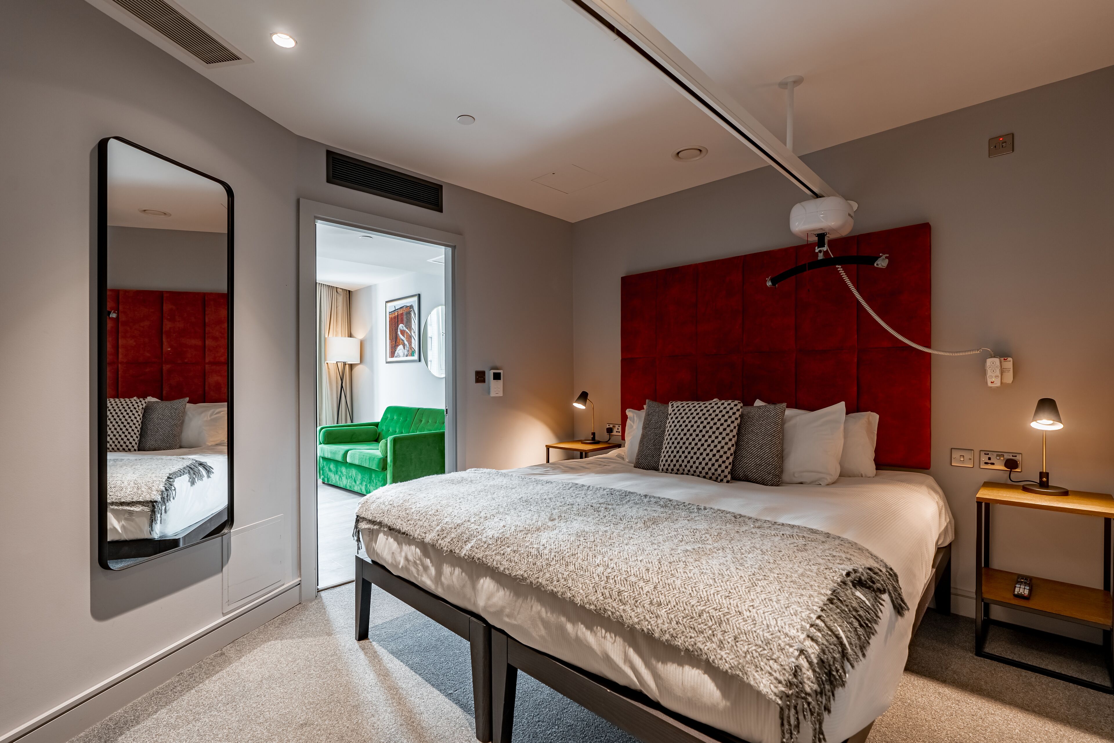 A modern hotel room with a large bed, a red headboard, a mirror, a bedside table with a lamp, and a view into a green sitting area.