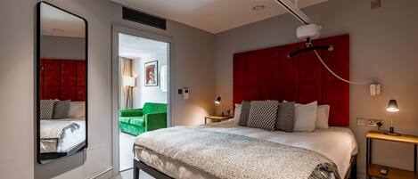 A modern hotel room with a large bed, a red headboard, a mirror, a bedside table with a lamp, and a view into a green sitting area.