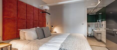 Une chambre d’hôtel moderne avec un grand lit, un mur d’accent rouge, un petit bureau et une salle de bain visible à travers une porte ouverte.
