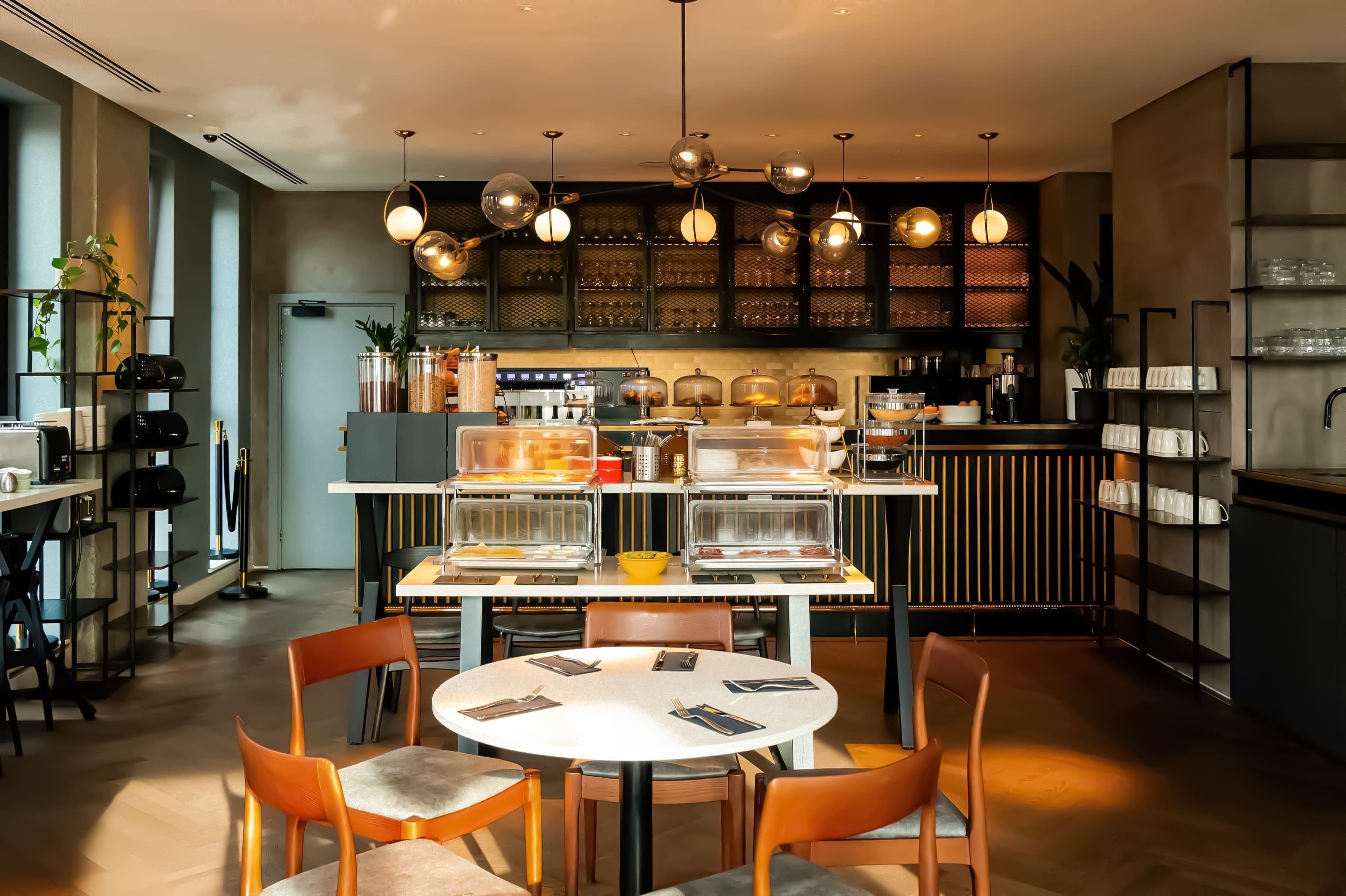 A modern cafe with a central round table, orange chairs, and a coffee bar in the background.