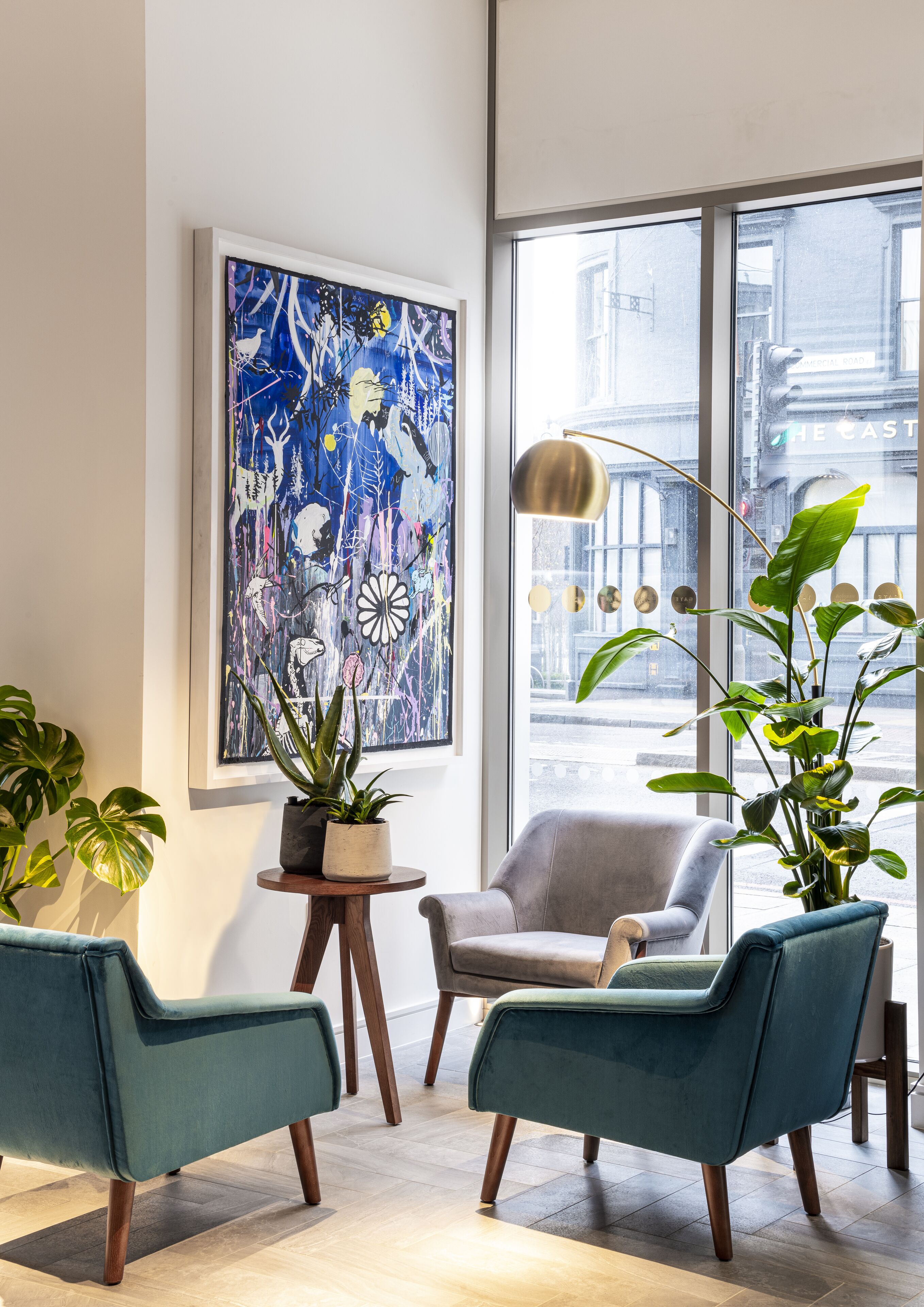 A modern living room with a large abstract painting, two teal armchairs, a grey sofa, and a tall floor lamp.