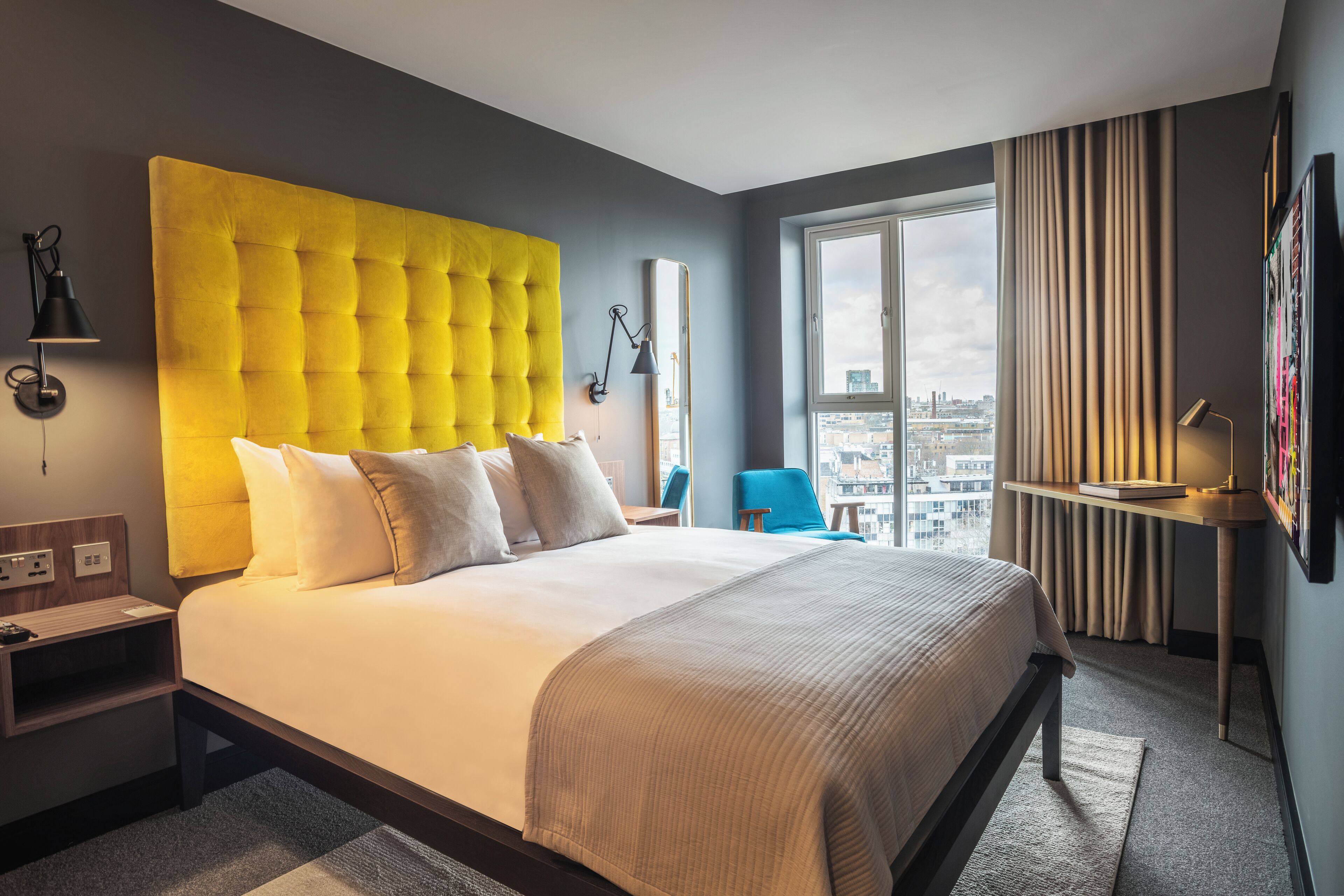 Une chambre d’hôtel avec un grand lit, une tête de lit jaune vif, une vue sur la ville par la fenêtre, un bureau avec une lampe et une chaise.