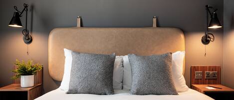 A neatly made bed with a headboard, two bedside lamps, a nightstand with a plant, and a wall socket.