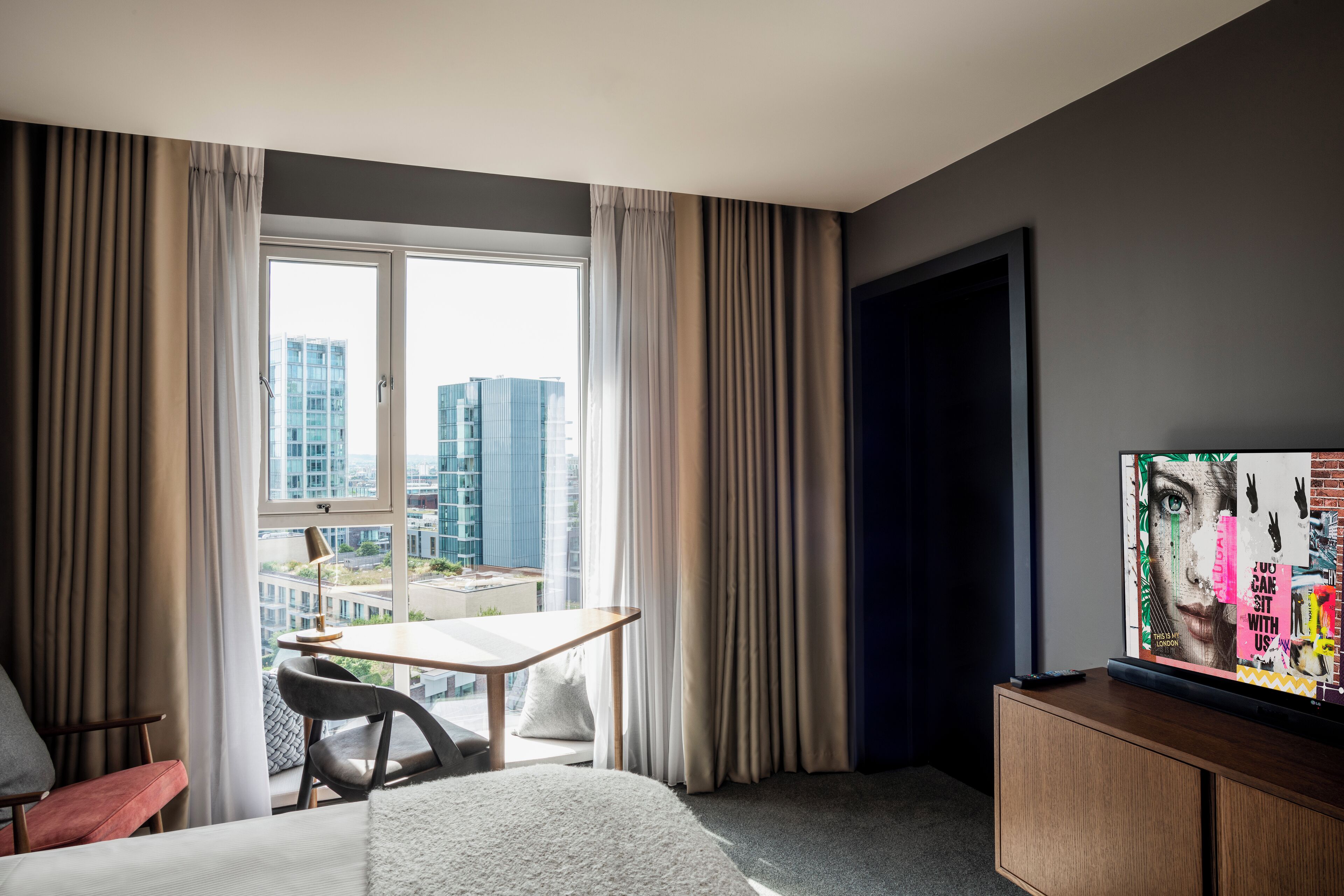 A hotel room with a large window, a desk, a bed, and a television.