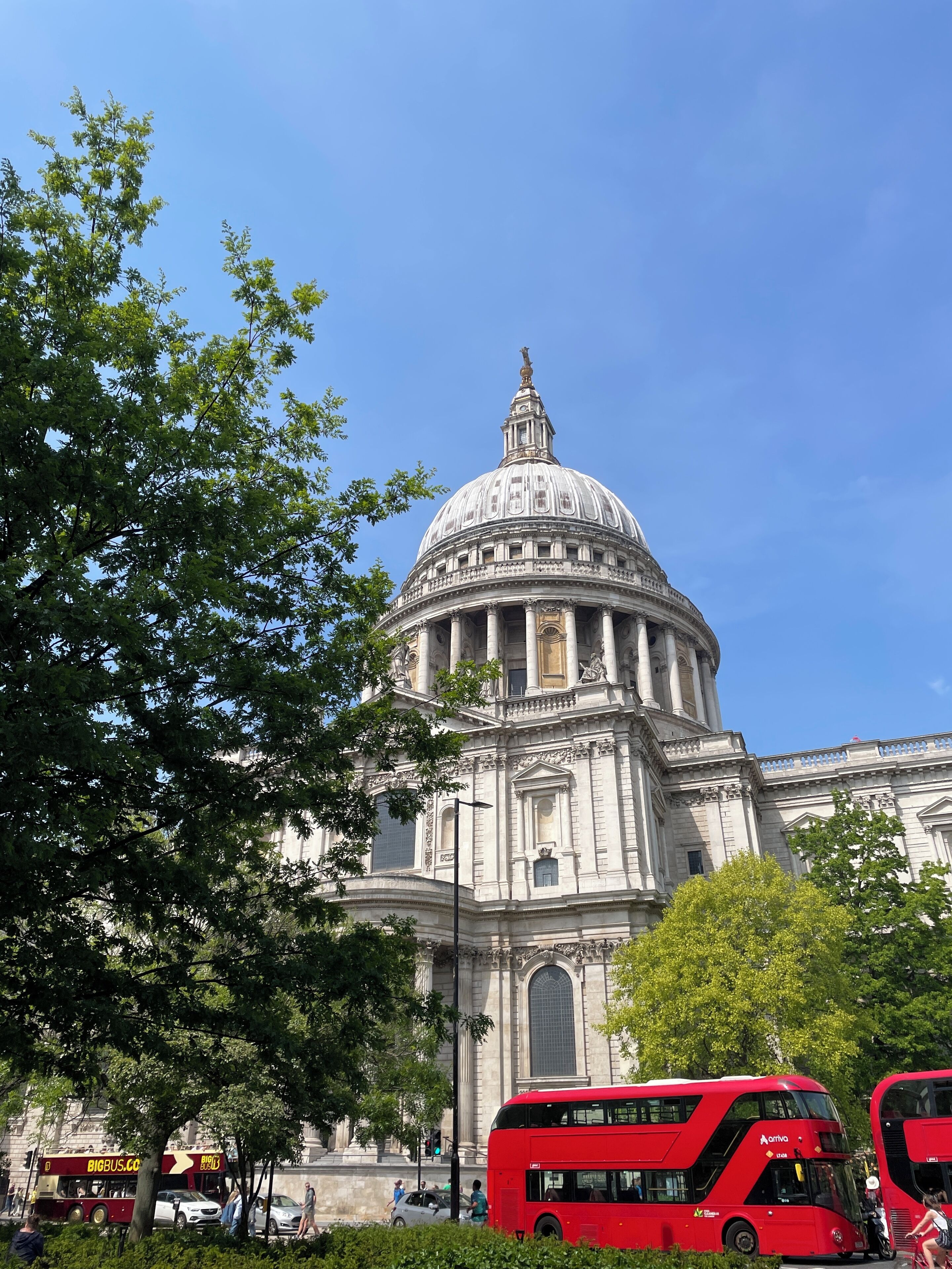 Un edifici gran de construcció clàssica, amb una cúpula destacada i columnes, envoltat d'arbres i autobusos vermells moderns.