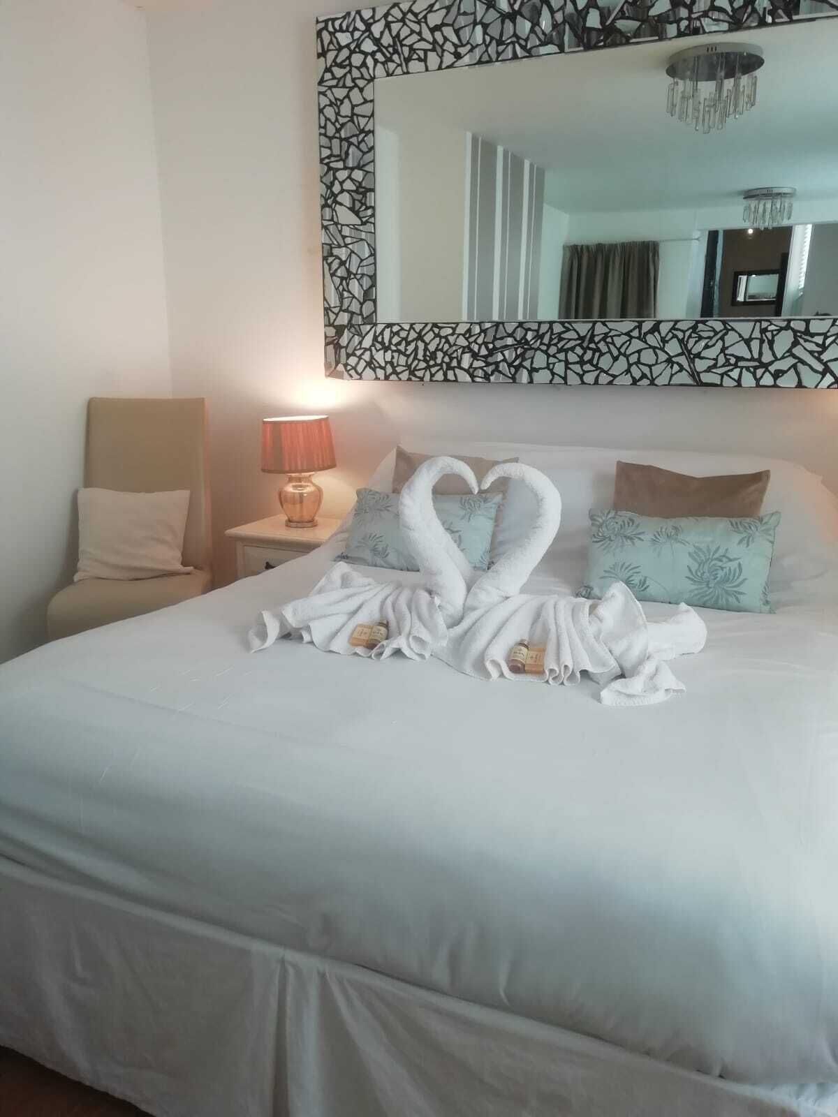 Cama bem arrumada com duas toalhas em formato de coração, um frasco de loção, uma luminária de cabeceira e uma cadeira em um quarto de hotel.