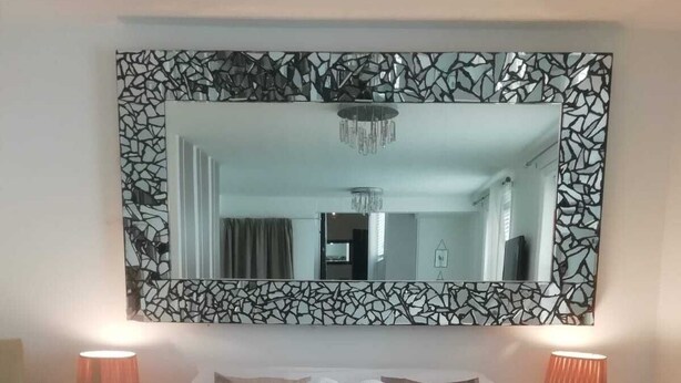 Cama bem arrumada com toalhas dispostas em formato de coração, um espelho grande com moldura de mosaico e duas luminárias de cabeceira.
