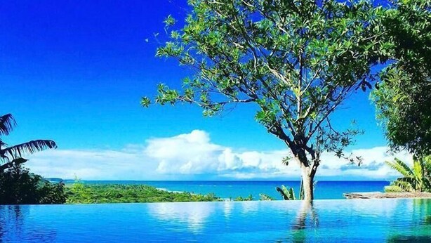 A clear blue infinity pool with a tree in the center, surrounded by lush greenery and a view of the ocean. - Pousada Segredos de Trancoso (Porto Seguro)