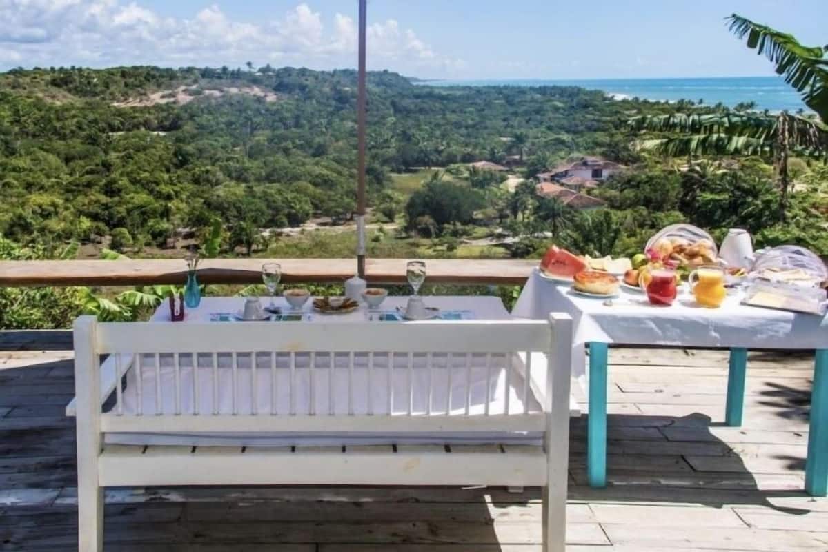Eine Holzterrasse mit einer weißen Bank und einem Tisch, der für eine Mahlzeit gedeckt ist, mit Blick auf eine üppige Landschaft und das Meer.