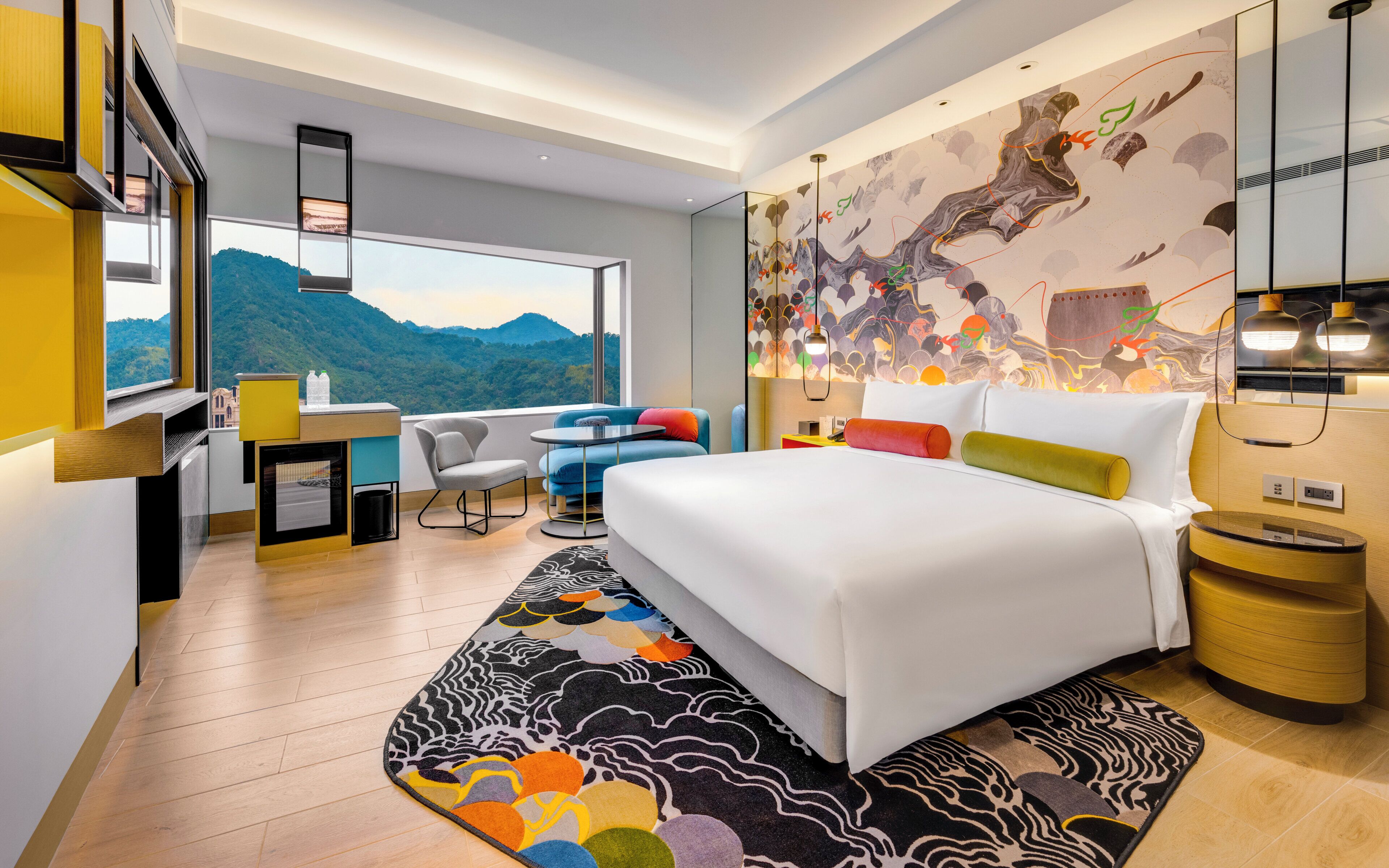 A modern hotel room with a large bed, a desk, a chair, and a view of the mountains.