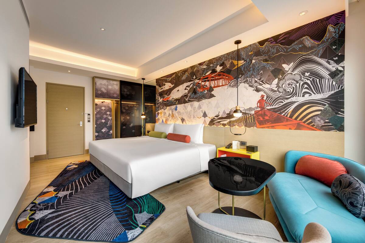 A modern hotel room with a large bed, a sofa, a TV, and a mural.