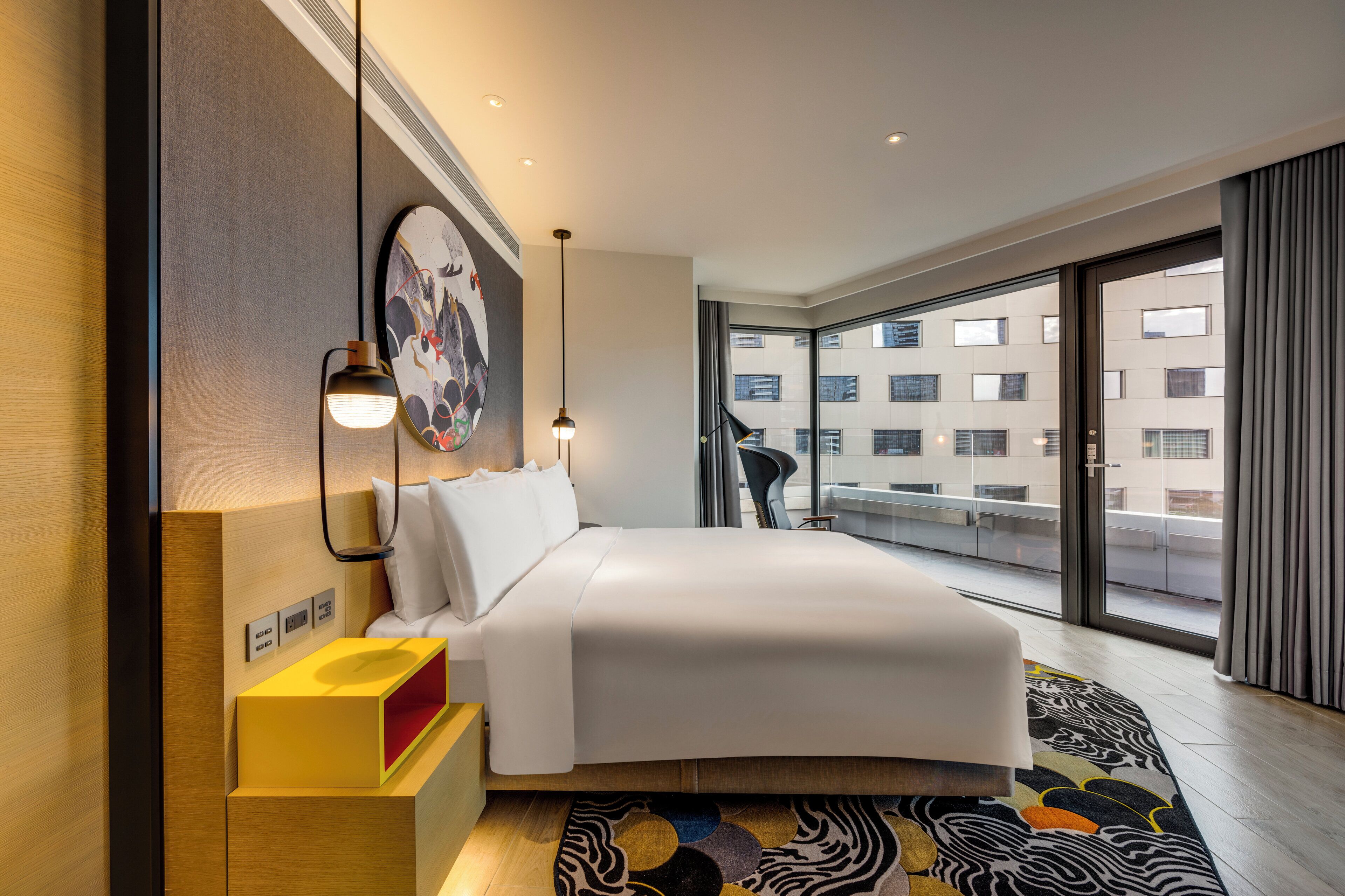 A modern hotel room with a large bed, a bedside table, a wall-mounted artwork, and a view of the city through large windows.