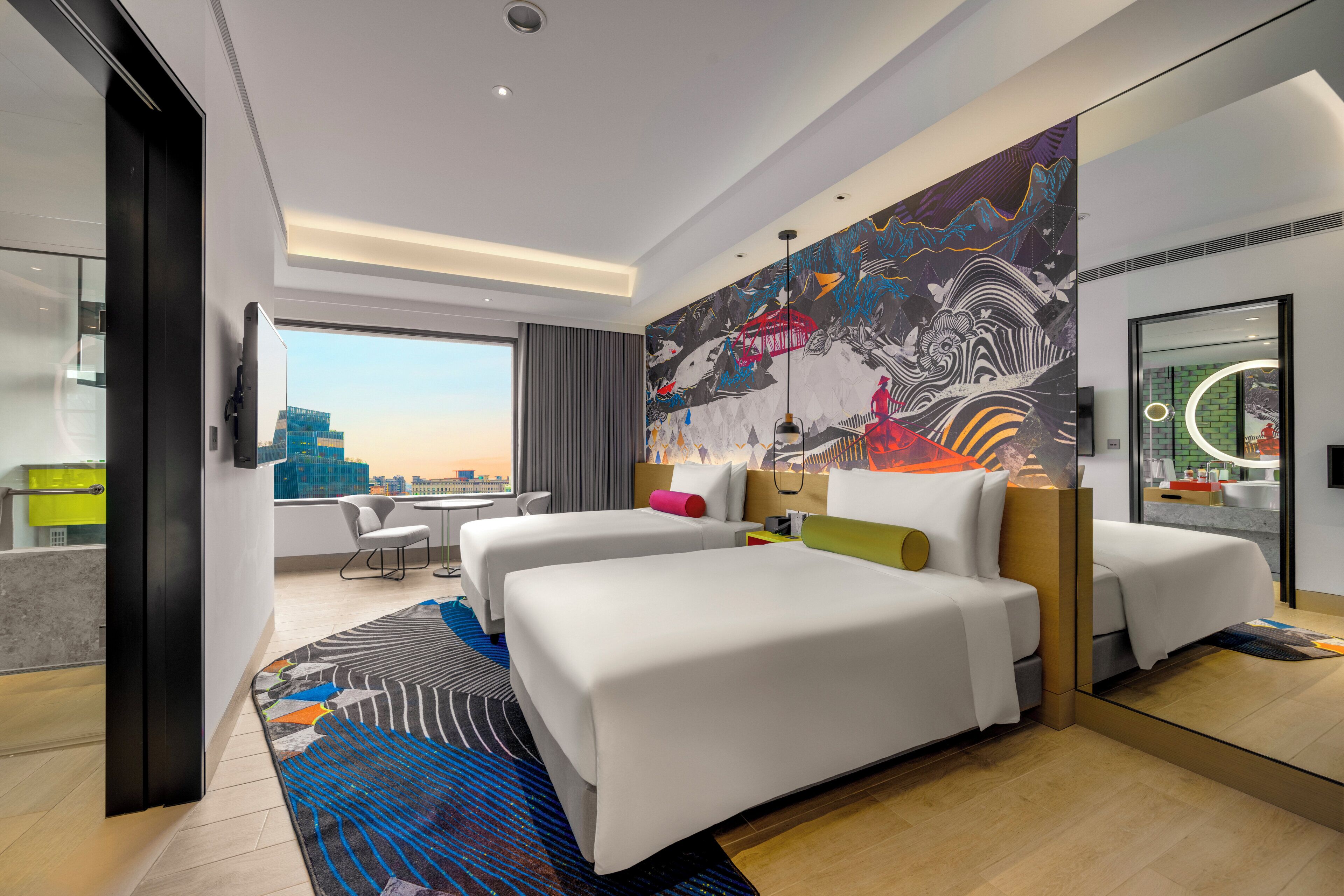 A modern hotel room with a large bed, a desk, and a view of the cityscape.
