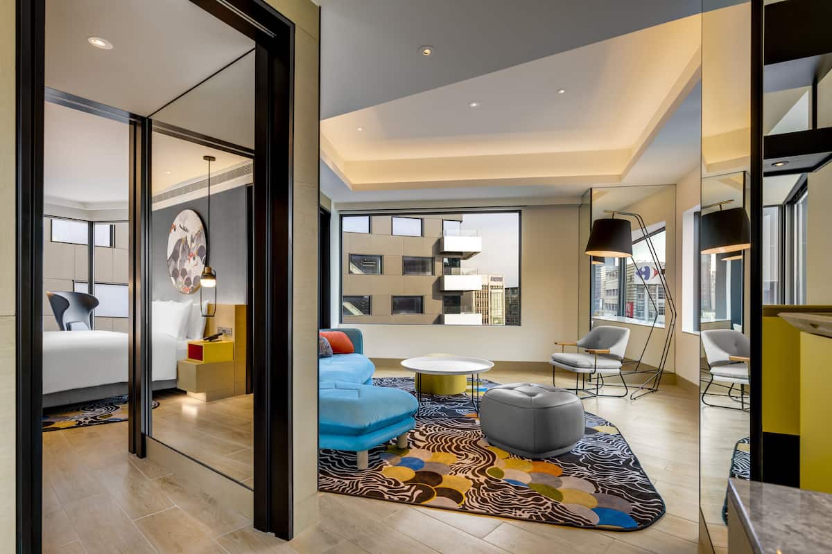 A modern hotel room with a large bed, a blue sofa, a gray ottoman, and a patterned rug.