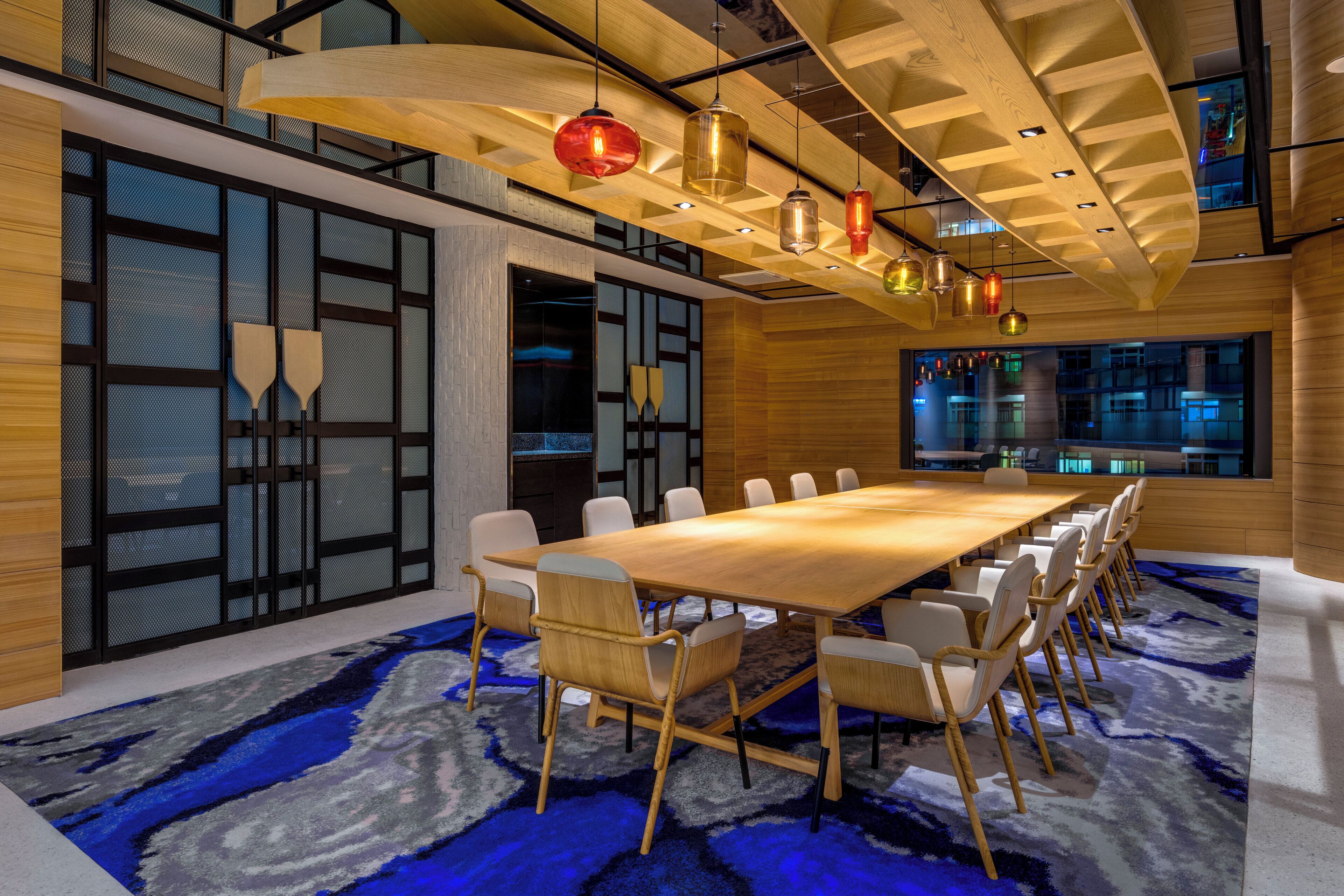 A conference room with a long wooden table, white chairs, and a blue patterned carpet.
