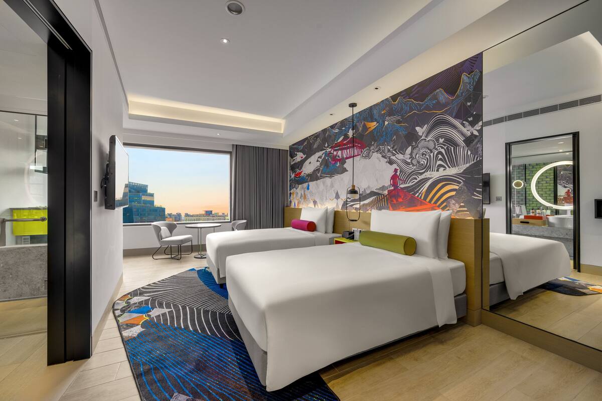 A modern hotel room with two beds, a large mural, a city view, and a glass partition.