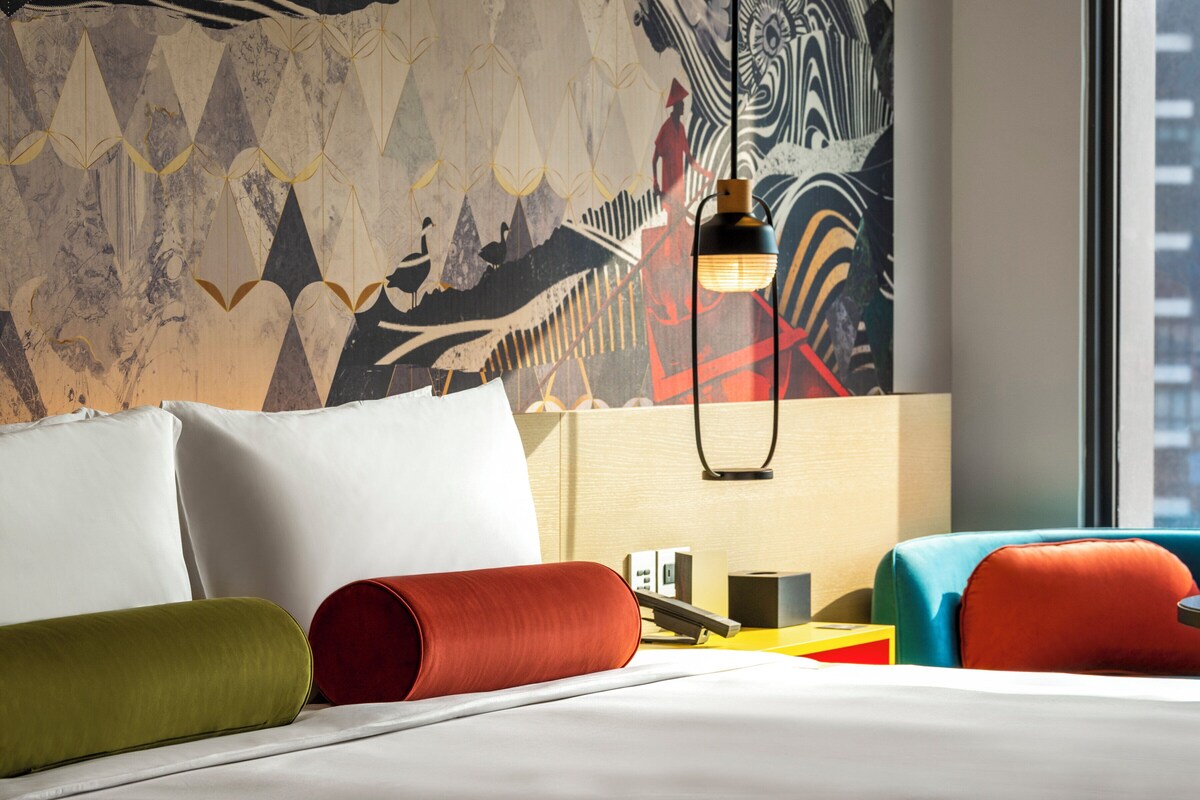 Ein Hotelzimmer mit einem Bett, Kissen, einer Lampe und einer Wand mit Kunstwerken.