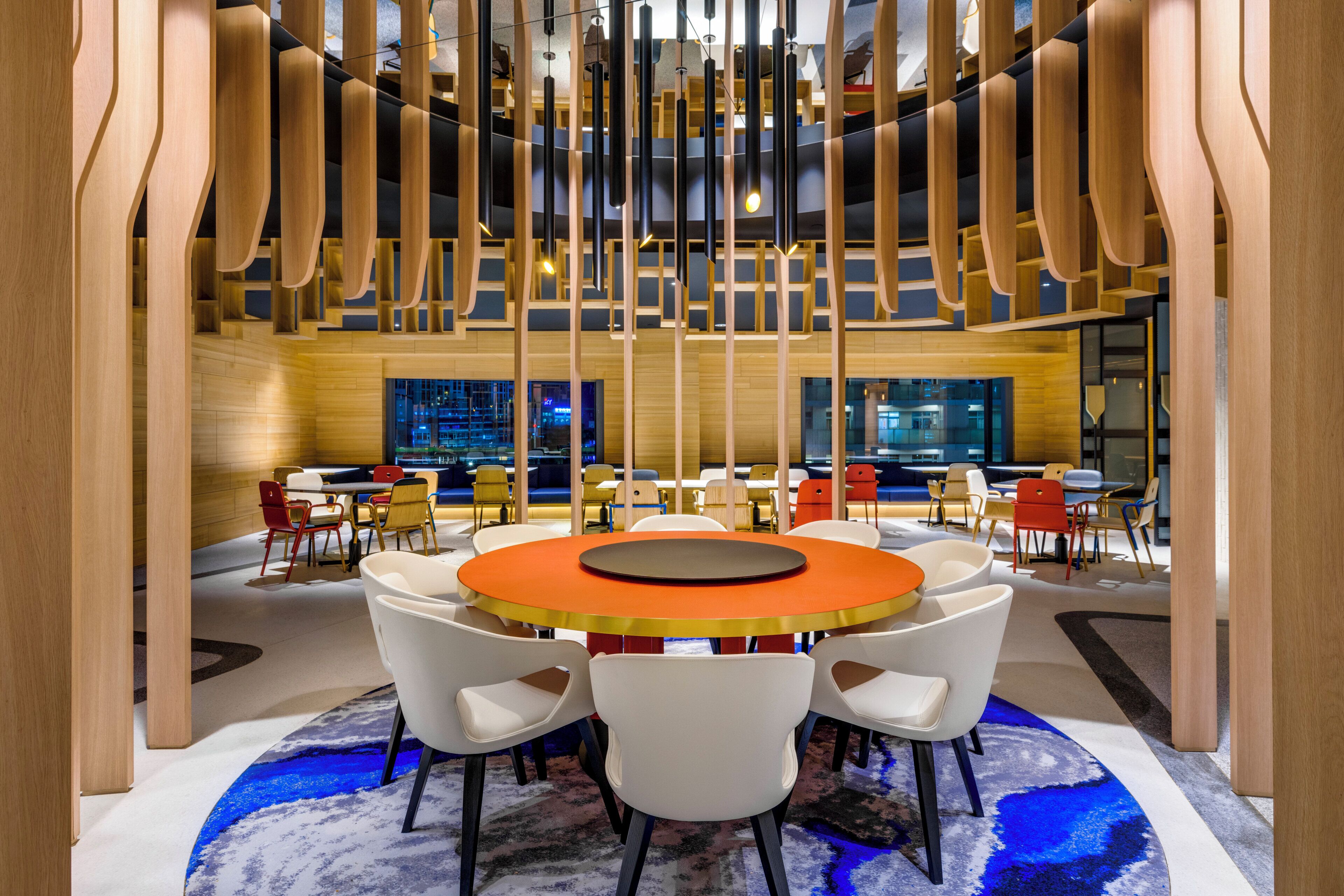 A modern interior with a central round table surrounded by white chairs, wooden accents, and a patterned carpet.