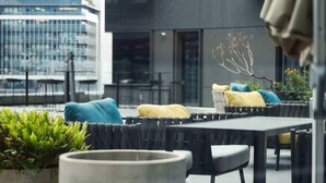 Un espace extérieur aménagé avec des meubles modernes, notamment une table et des chaises avec des coussins, ainsi qu’un bac à fleurs orné de plantes vertes.