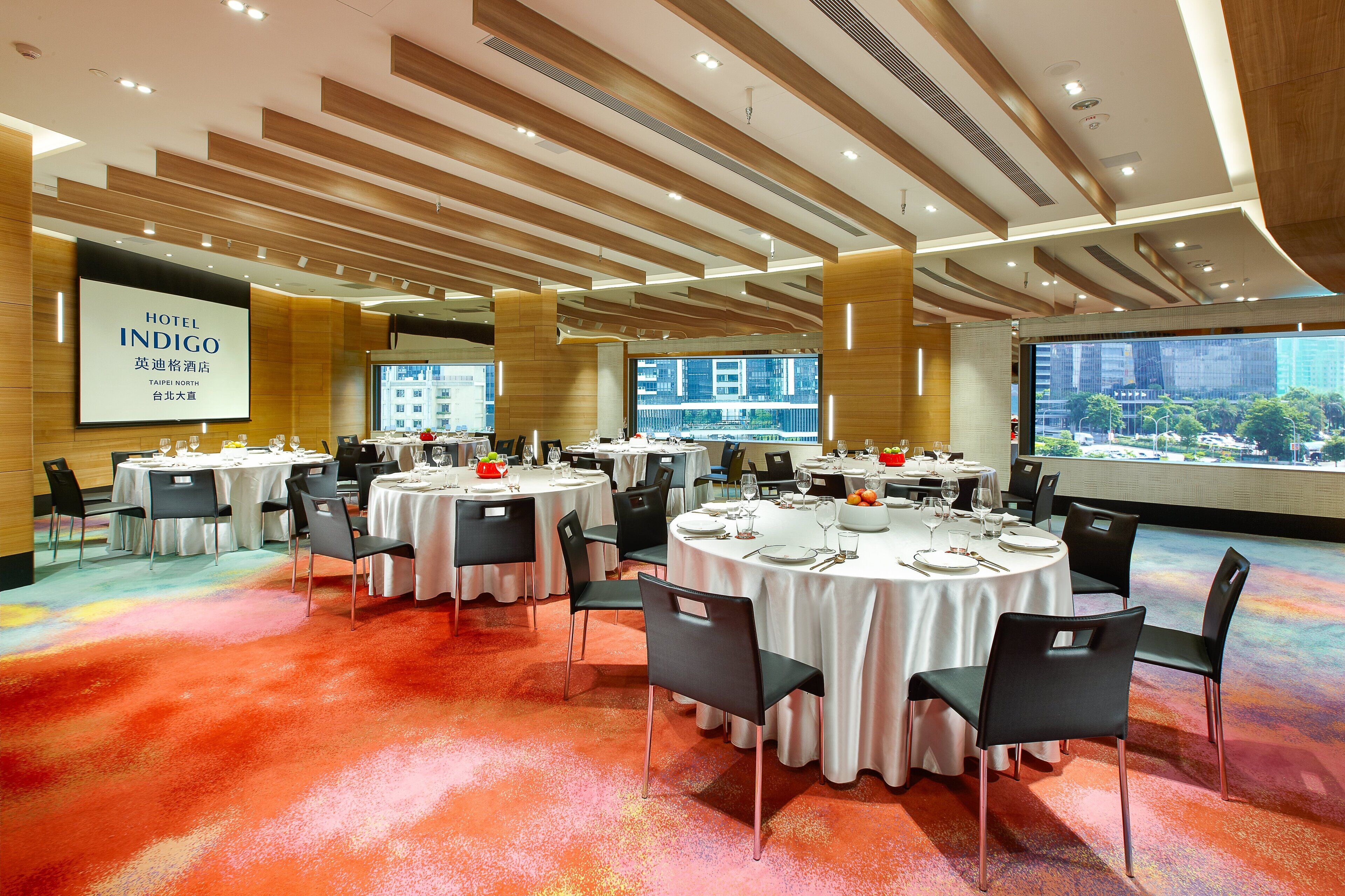 A hotel conference room with multiple round tables, black chairs, and a large screen displaying the hotel name.