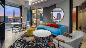 Un salon moderne avec un canapé bleu, un miroir rond et une vue sur les toits d’une ville au coucher du soleil.
