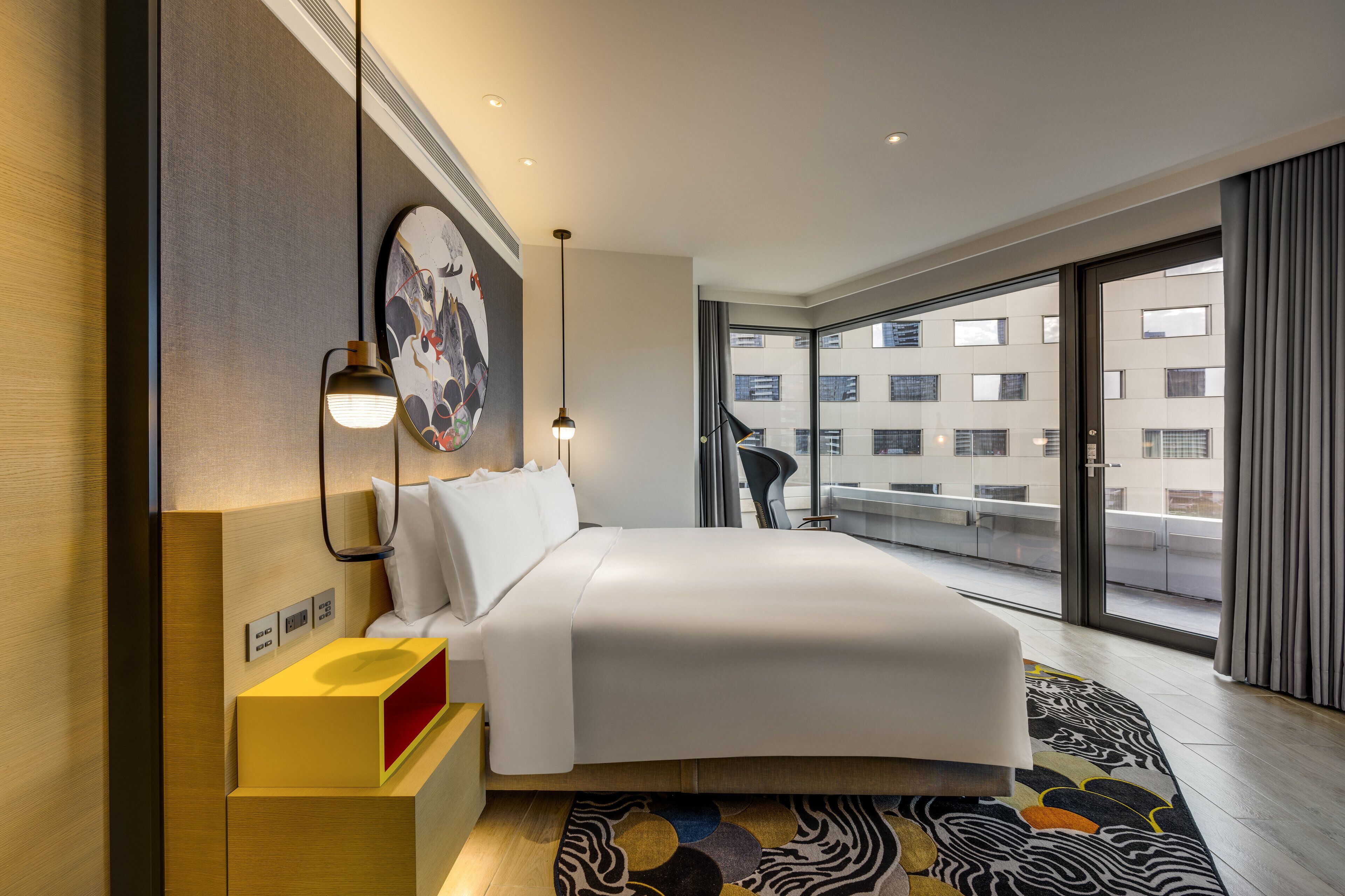 A modern hotel room with a large bed, a bedside table with a lamp, a wall-mounted artwork, and a view of the city through large windows.