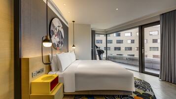A modern hotel room with a large bed, a bedside table with a lamp, a wall-mounted artwork, and a view of the city through large windows.
