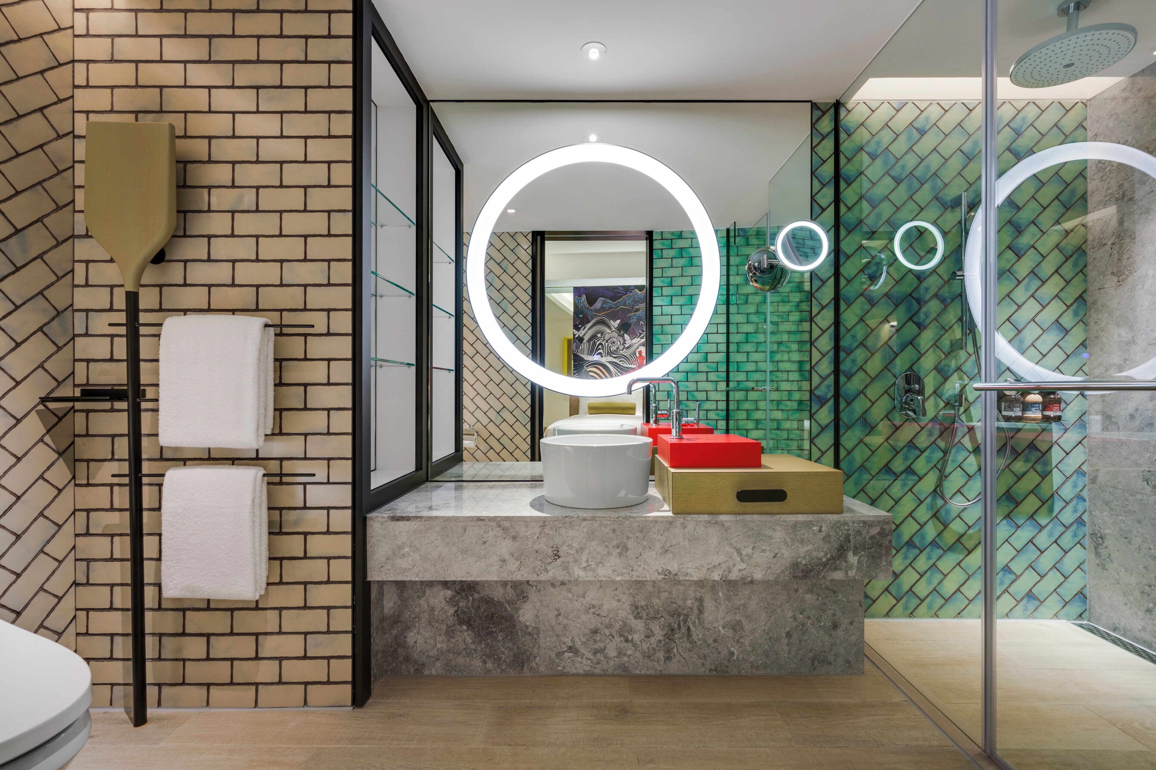 Een moderne badkamer met een bakstenen muur, een ronde spiegel met verlichting, een wit wastafel en een glazen douchecabine.