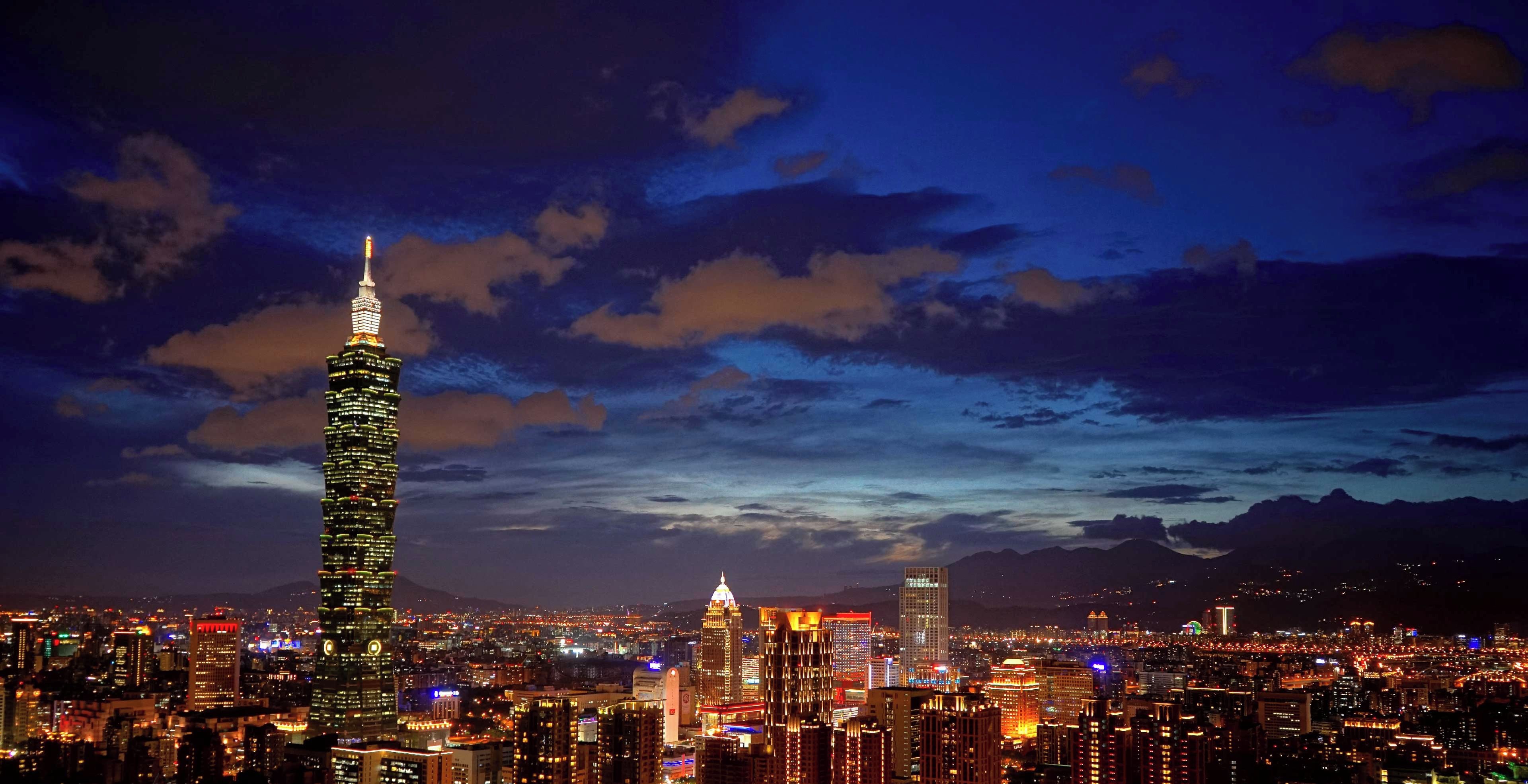 A cityscape at night with illuminated skyscrapers, including a prominent tower with a spire.