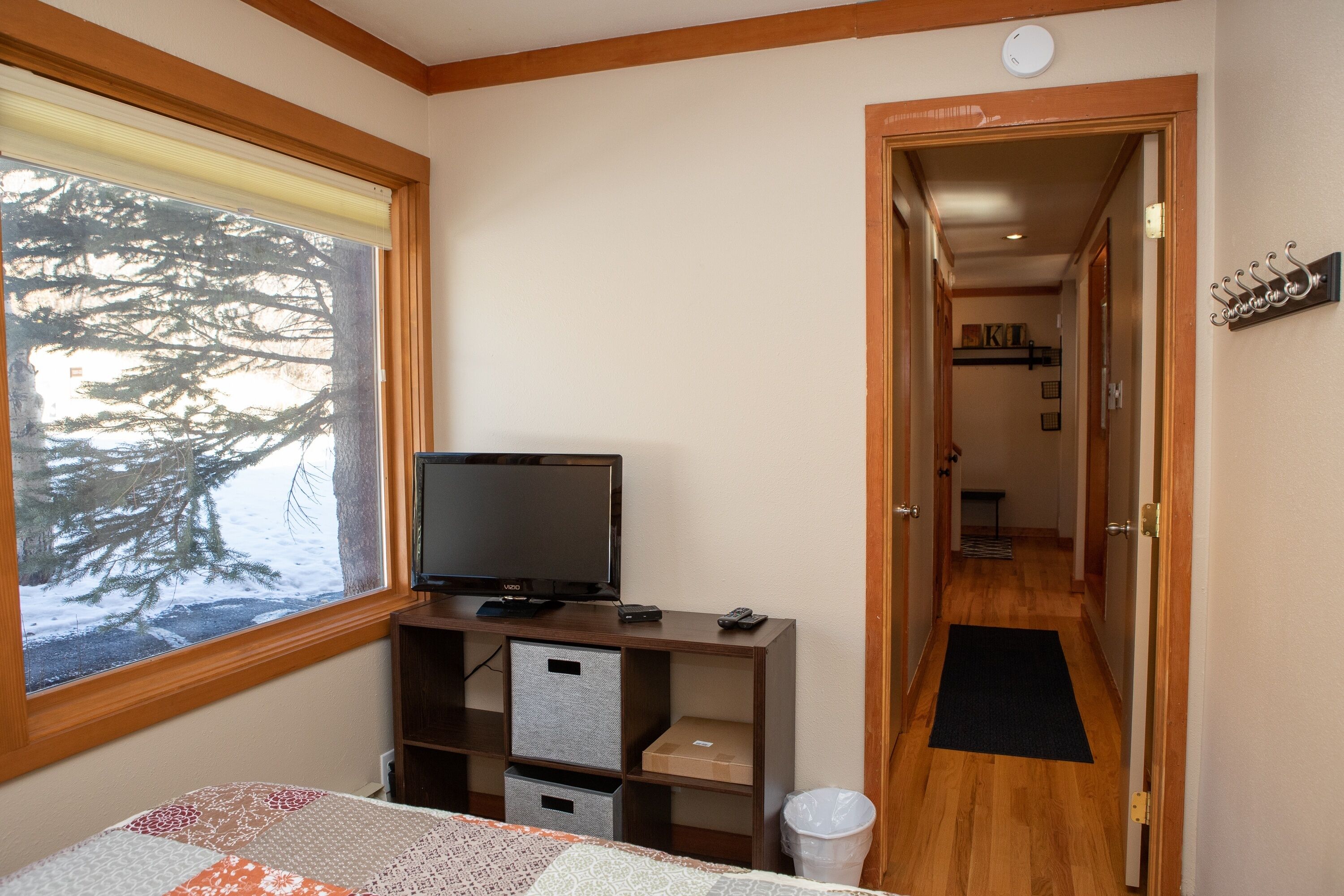 A room with a bed, a TV on a wooden stand, a window with a view of trees, and a door leading to another room.