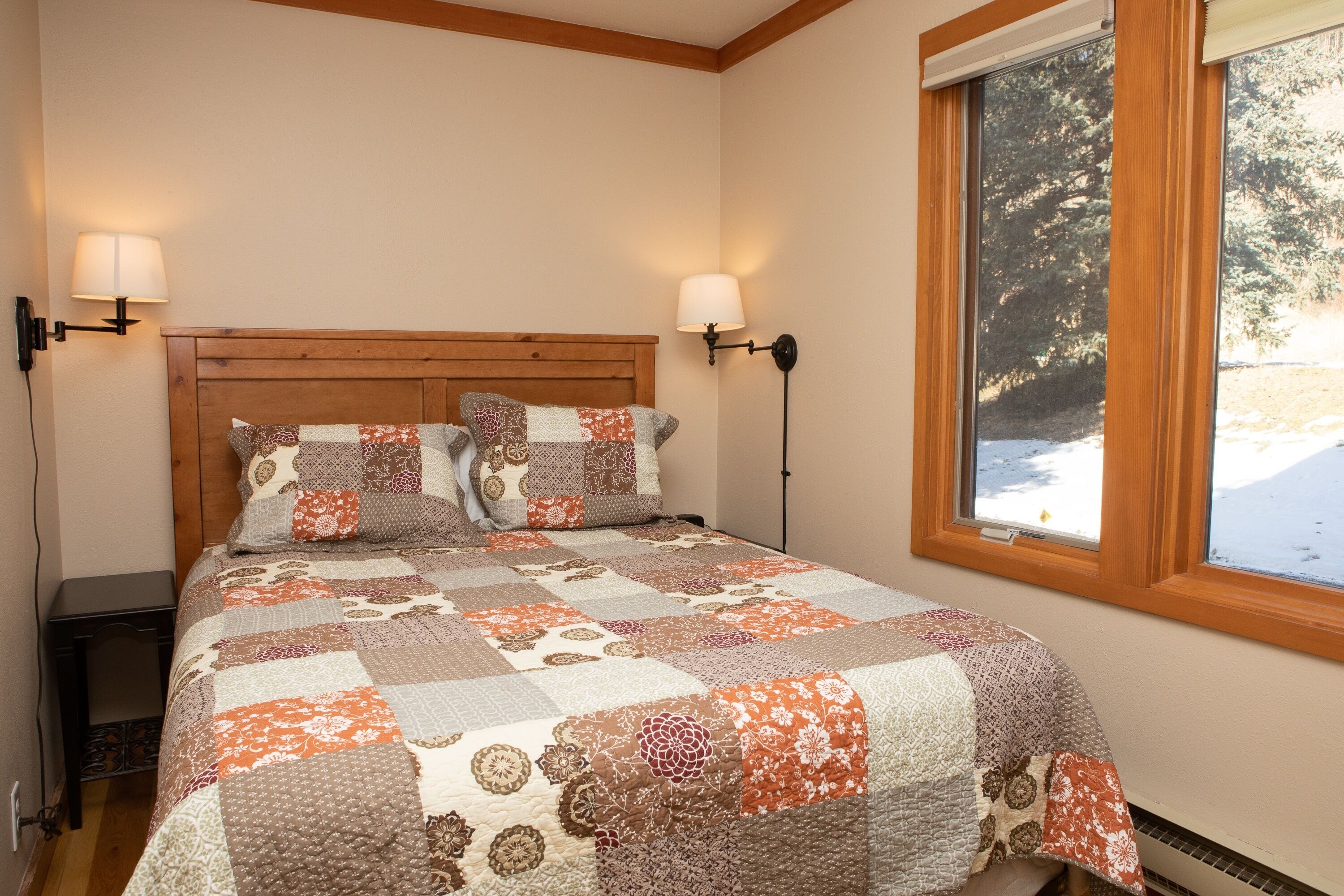 A bedroom with a wooden headboard, a quilted bedspread, two bedside lamps, and a window with a view of trees.