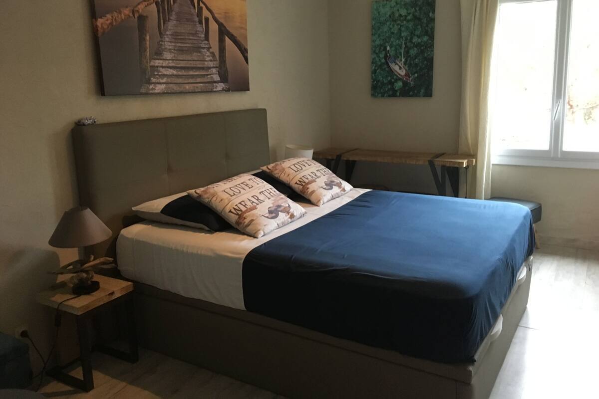 A bedroom with a bed, a nightstand, a lamp, a painting of a bridge, and a window with curtains.