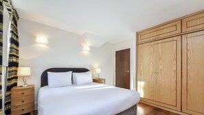 A neatly made bed with white linens, a wooden nightstand with a lamp, and a large wooden wardrobe. - Two bedroom apartment with sofa bed (London)