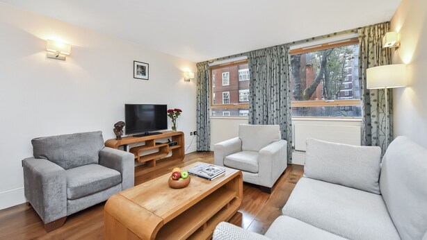 A modern living room with a grey sofa, a white armchair, a wooden coffee table with a fruit bowl, and a flat-screen TV on a wooden stand. - Two bedroom apartment with sofa bed (London)
