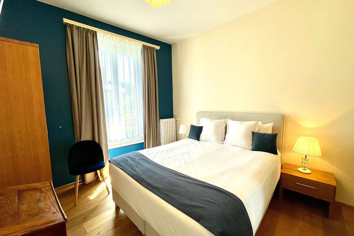 Une chambre d’hôtel avec un grand lit, un bureau, une chaise, une lampe et une fenêtre avec des rideaux.