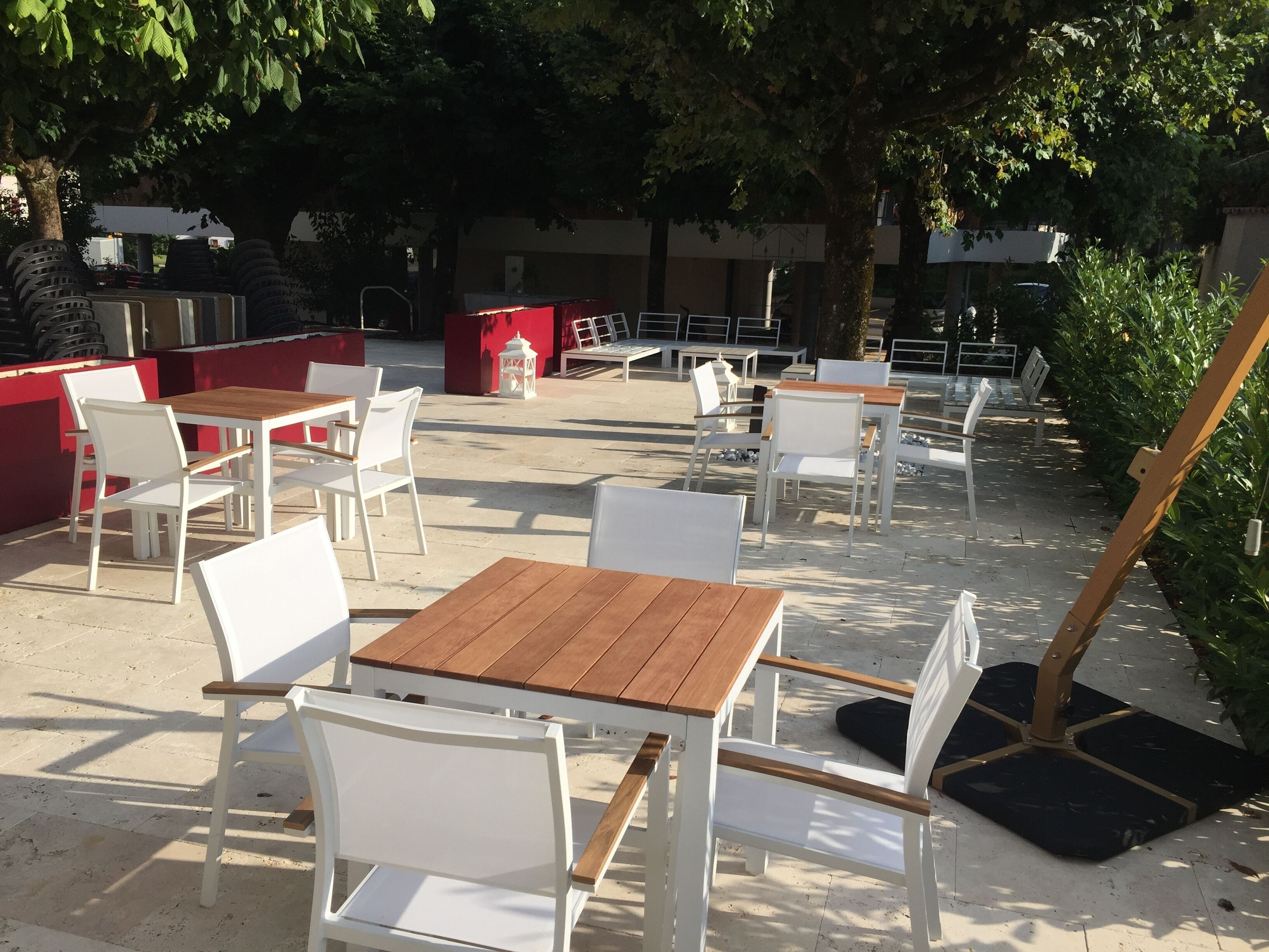 An outdoor seating area with white chairs and wooden tables, surrounded by trees and a red wall.