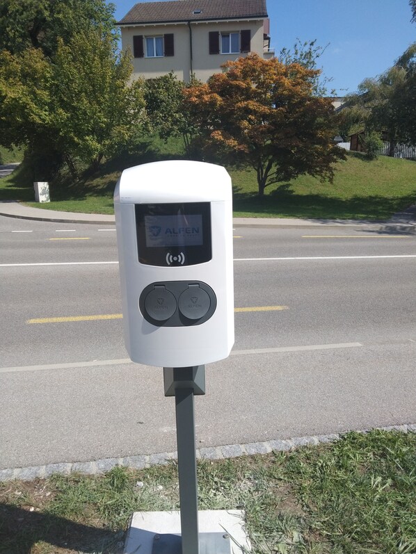 A parking meter with a display and two charging ports, located on a street with a house and trees in the background. - Hotel La Belle Croix (Romont)