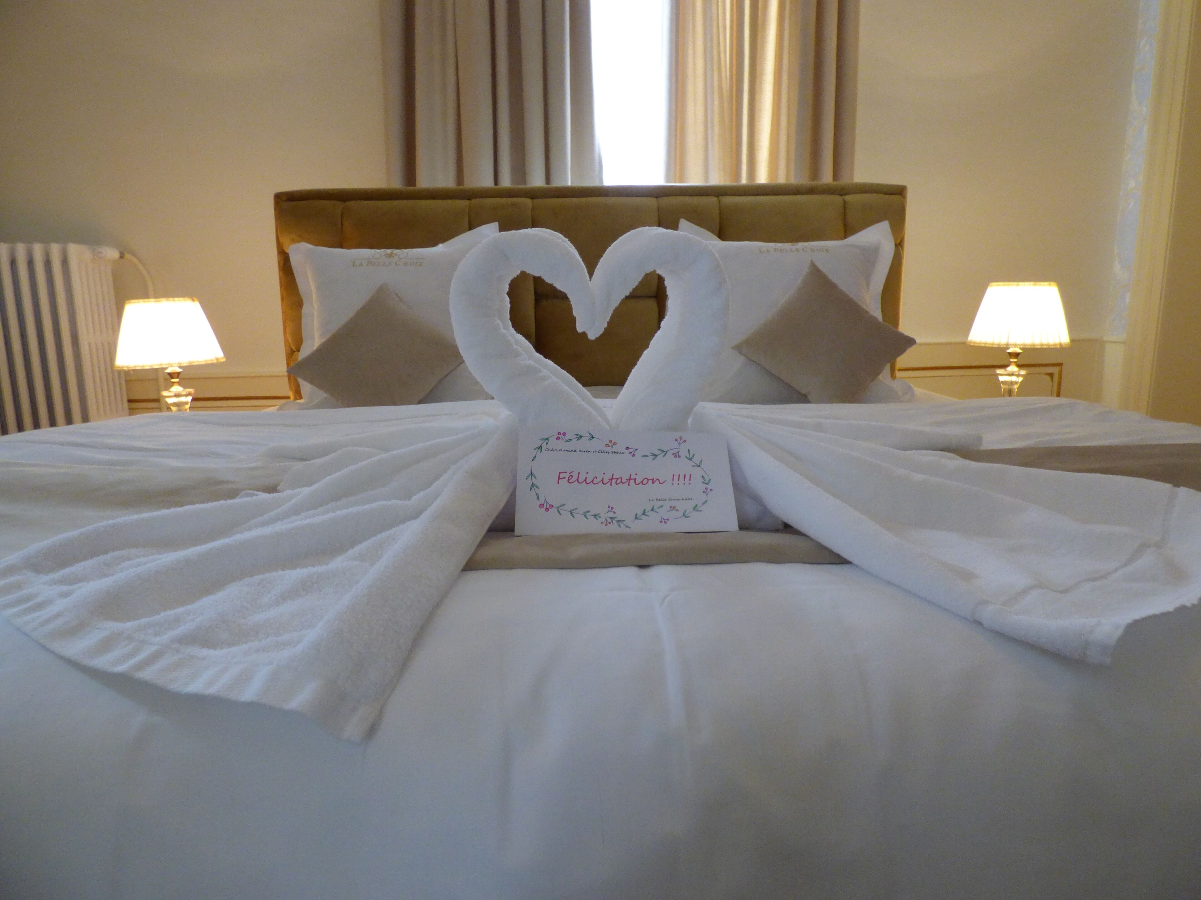 A neatly made bed with a heart-shaped towel arrangement and a card that reads "Felicitation!!!".