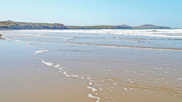 Ein Strand mit Wellen, Sand und sanften Hügeln im Hintergrund.