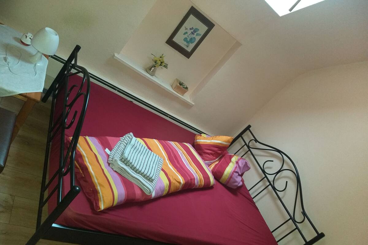 Ein Einzelbett mit roter Matratze, gestreifter Bettwäsche und einer zusammengelegten Decke. Darüber am Bett befindet sich ein kleines Regal mit einem Blumenstrauß.