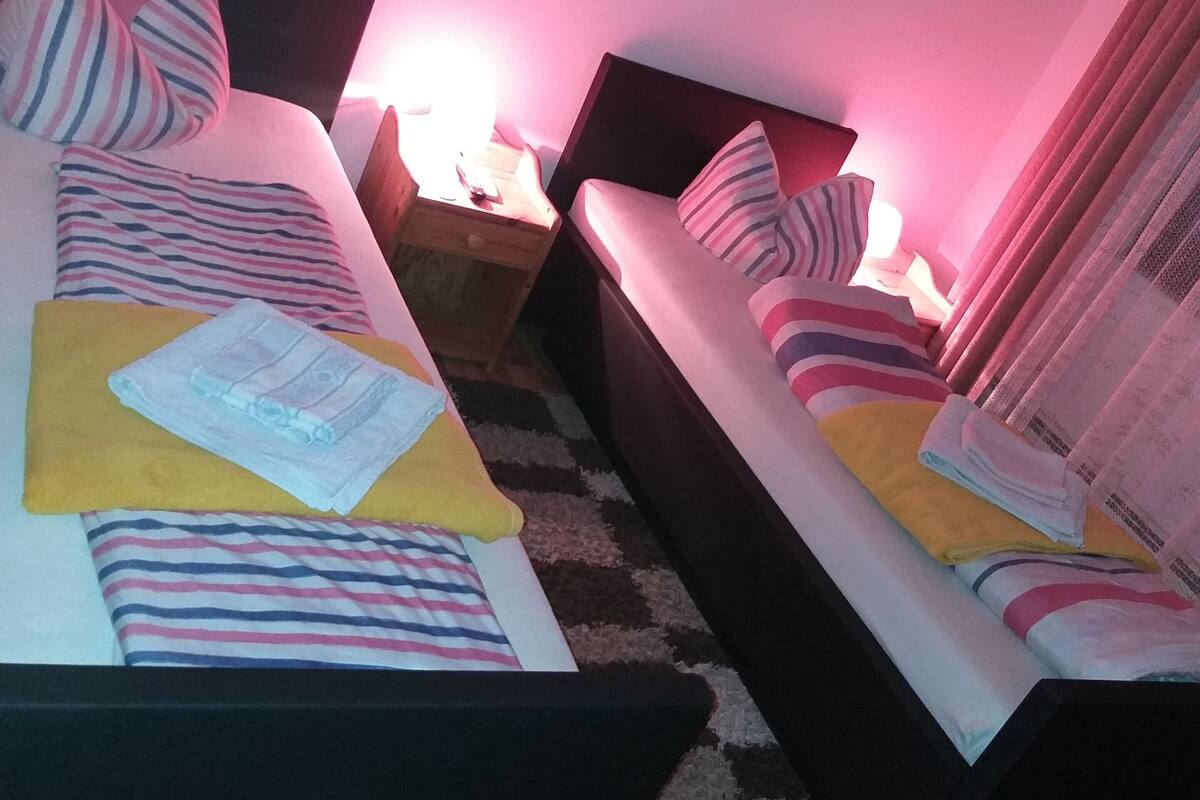 Zwei Einzelbetten mit gestreifter Bettwäsche, ein hölzernes Kopfstück, ein Nachttisch mit Lampe und ein kleiner Tisch mit Fernbedienung.