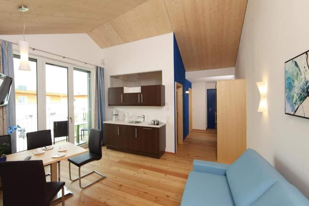 Un salon moderne avec un canapé bleu, un plancher en bois, un coin repas avec une table et des chaises, une cuisine avec des meubles sombres et un téléviseur fixé au mur.