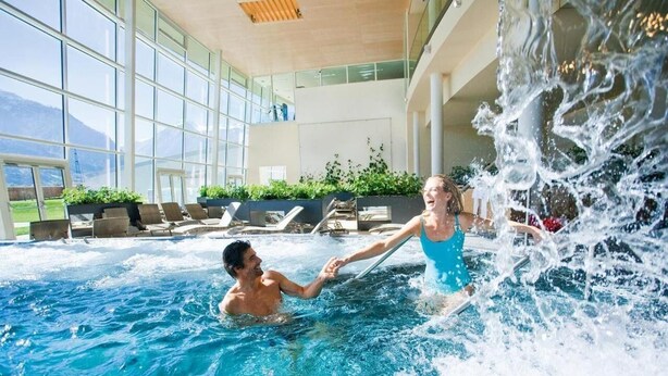 An indoor swimming pool with clear glass walls, a hot tub, and lounge chairs.